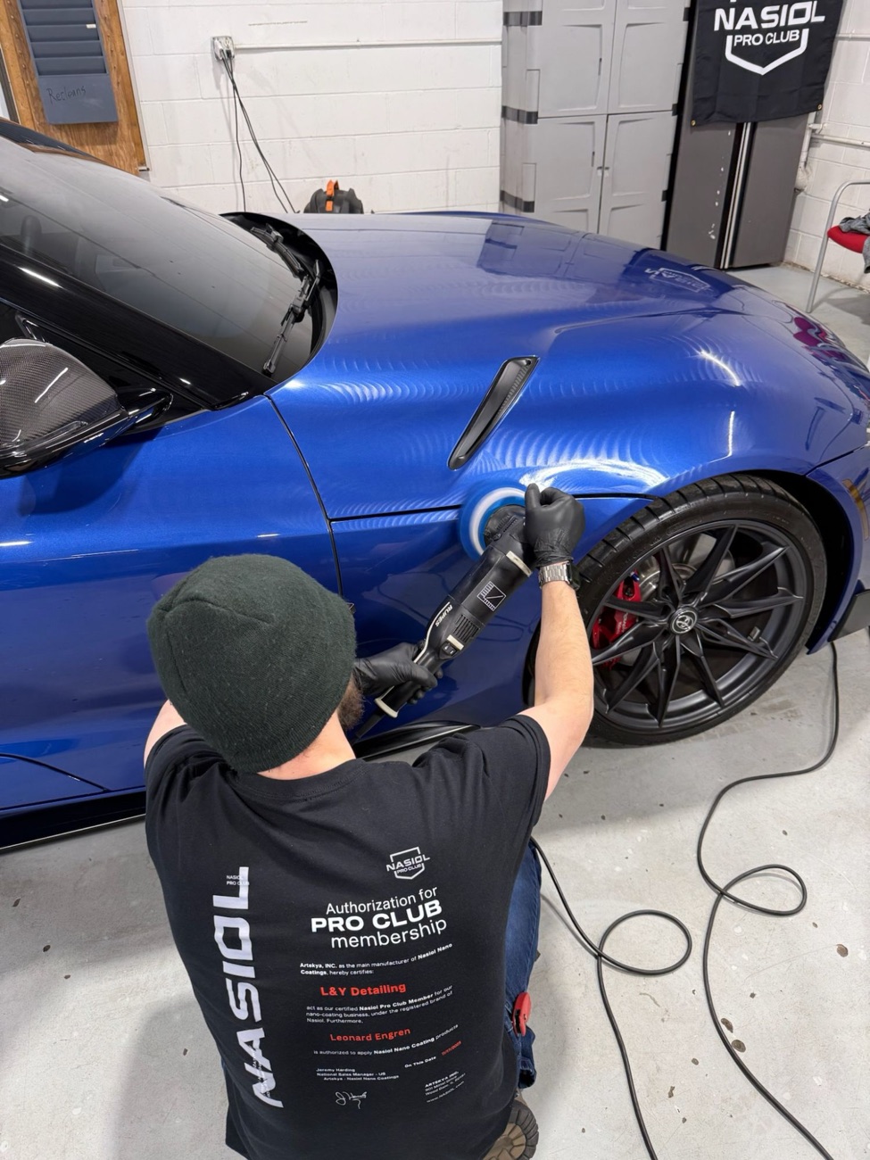 Leonard machine-polishing a blue Toyota Supra, wearing his Nasiol Pro Club authorization shirt